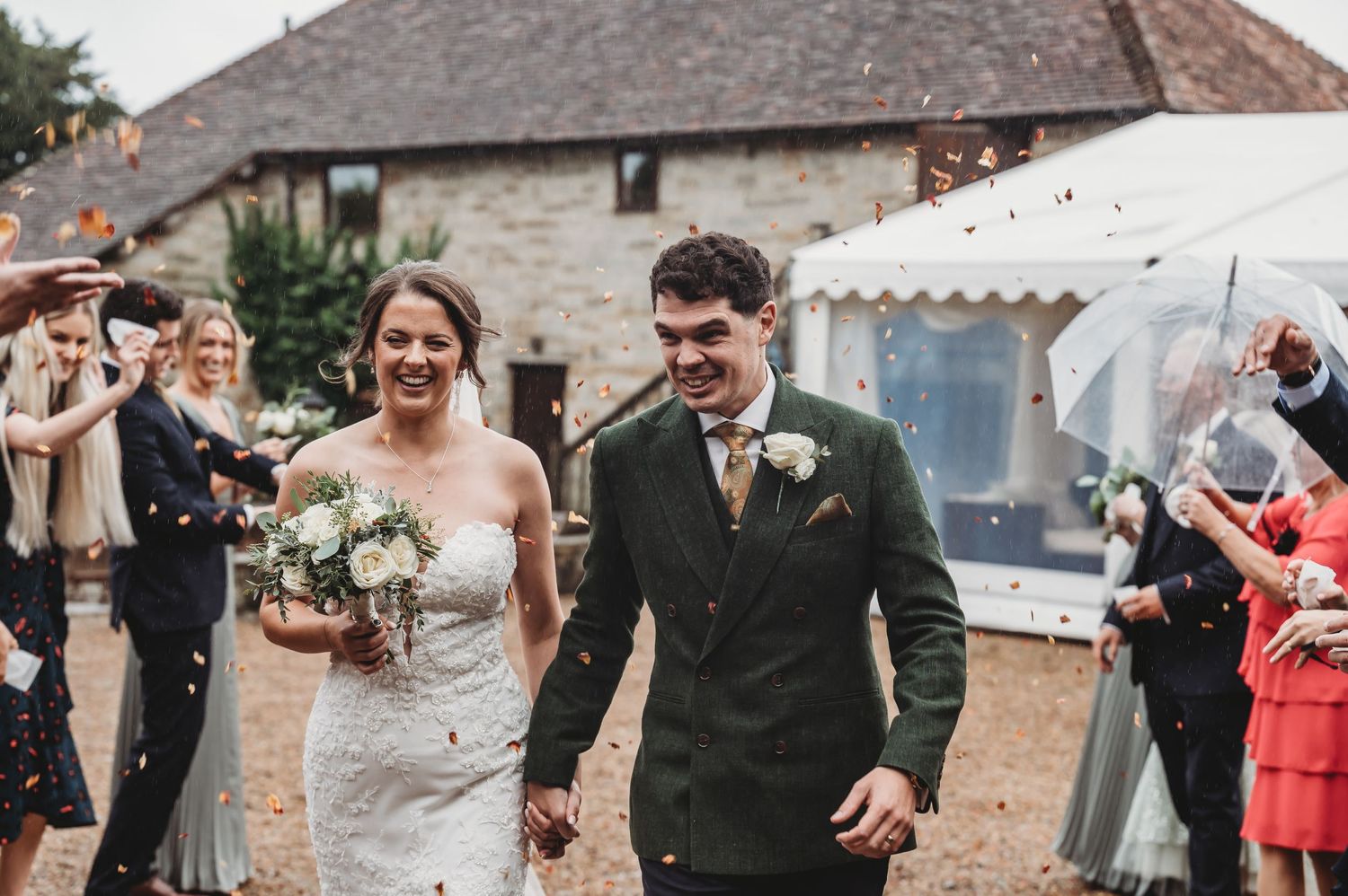 Joyful wedding couple walks through confetti shower outside stone building with guests celebrating.