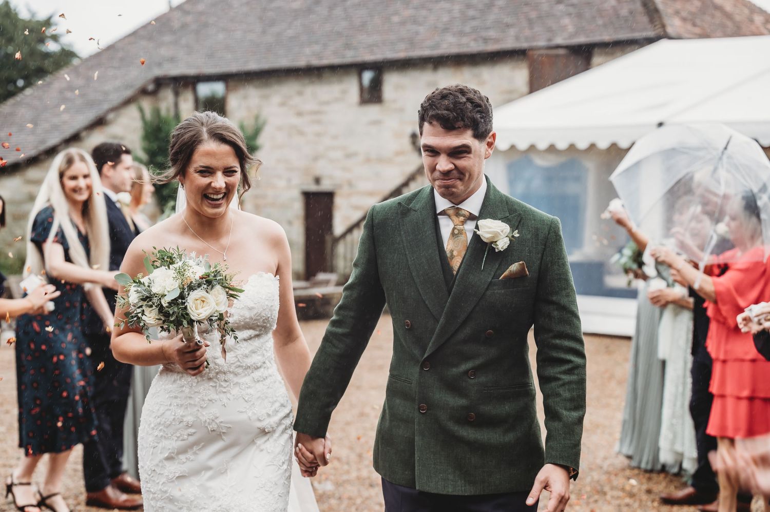 Wedding couple walks together as guests shower them with confetti near a stone building.
