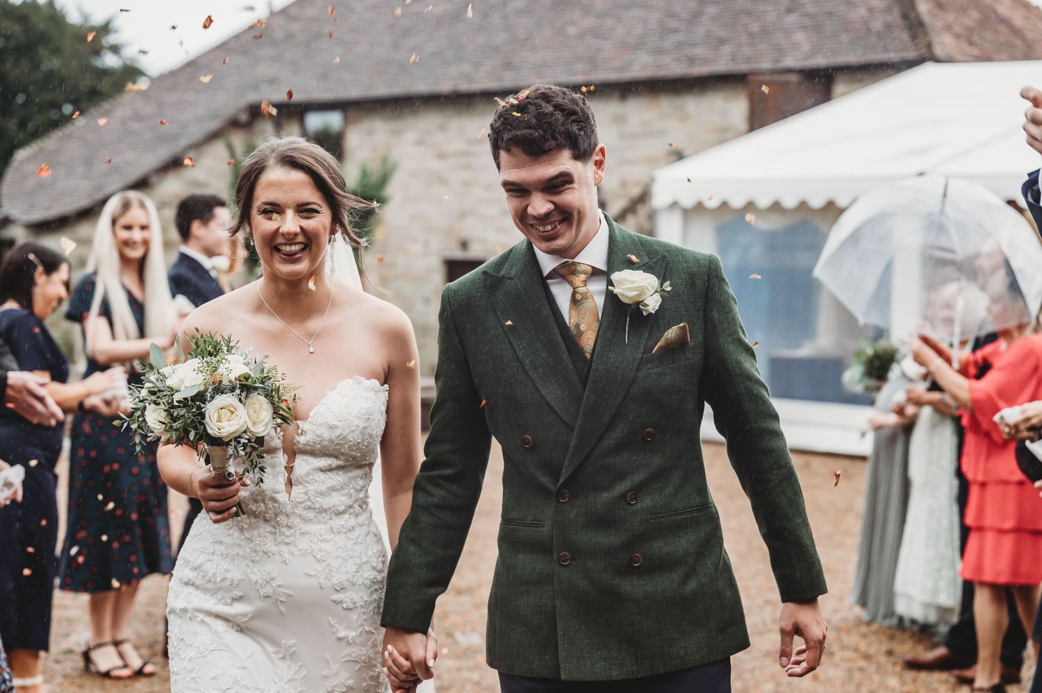 Newlyweds walk hand in hand while guests throw confetti outside a rustic stone building.