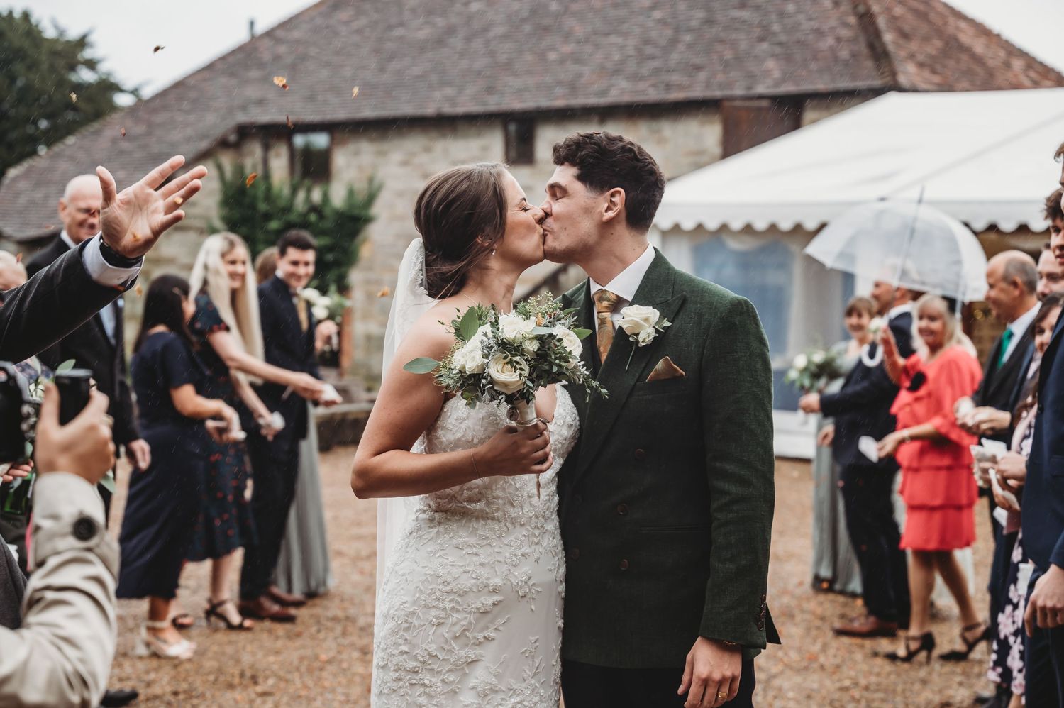 Wedding guests celebrate with umbrellas as newlyweds share a moment outside a stone building.