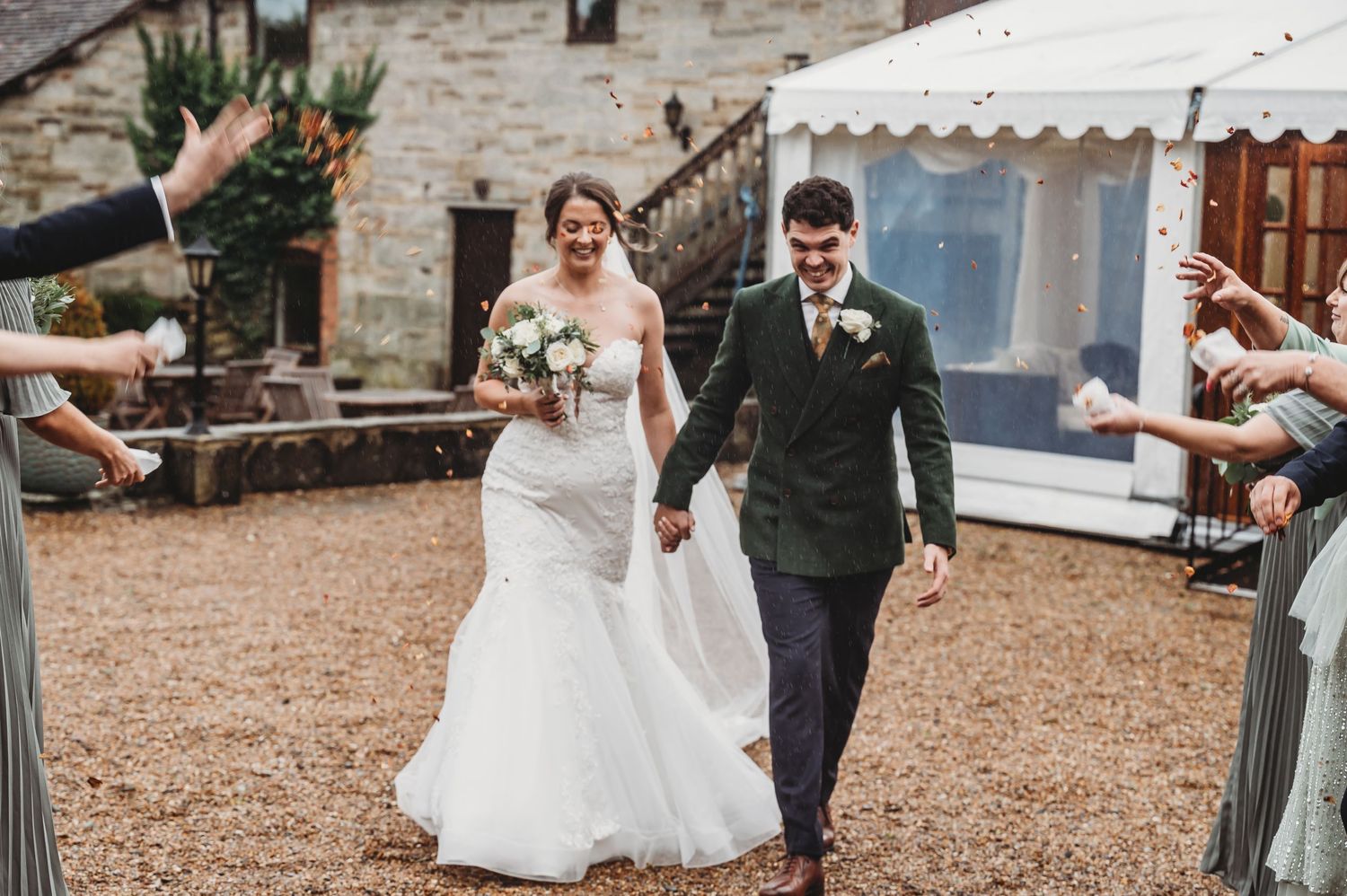 A couple walks hand in hand while guests celebrate their wedding exit at a rustic stone venue.