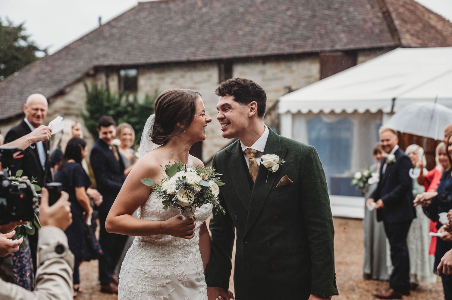 Newlyweds share a moment outside a stone building while wedding guests celebrate around them.