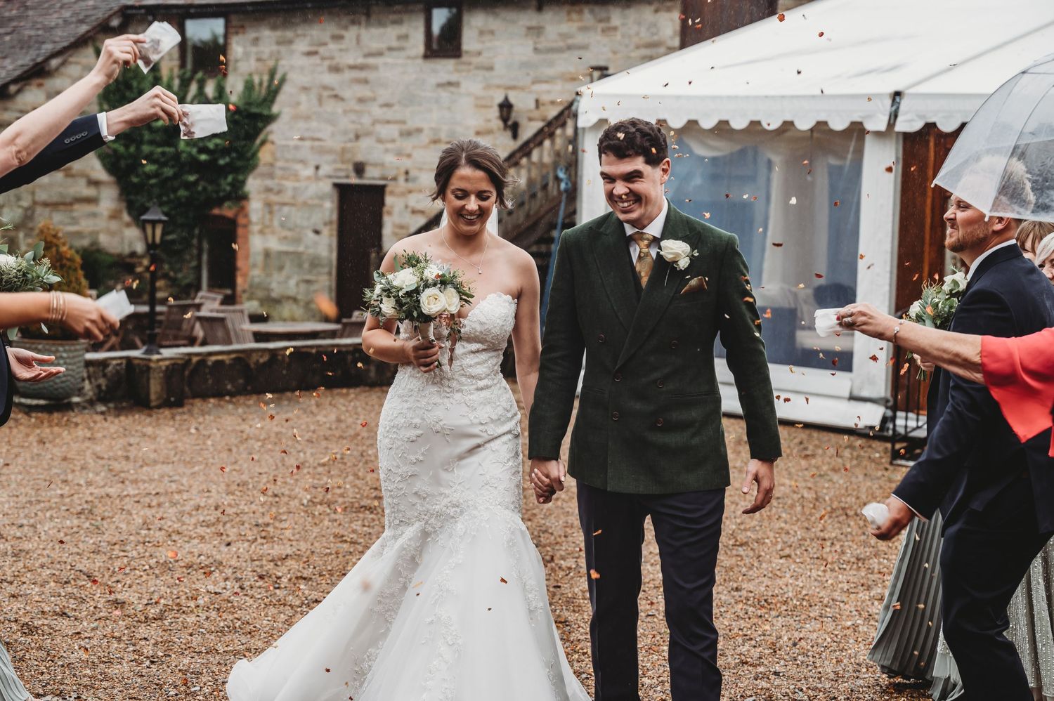 Wedding couple exits their ceremony while guests throw confetti outside a rustic stone venue.