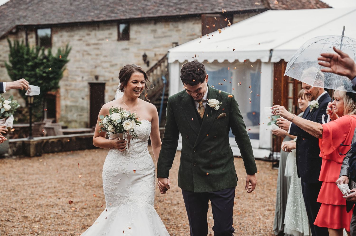 Couple in wedding attire walks past celebrating guests at rustic stone venue.