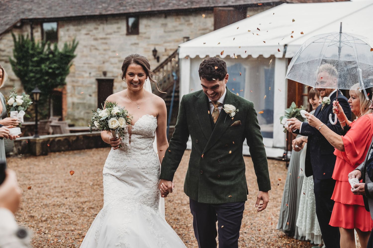 A wedding couple walks together on a gravel courtyard while guests shower them with confetti.