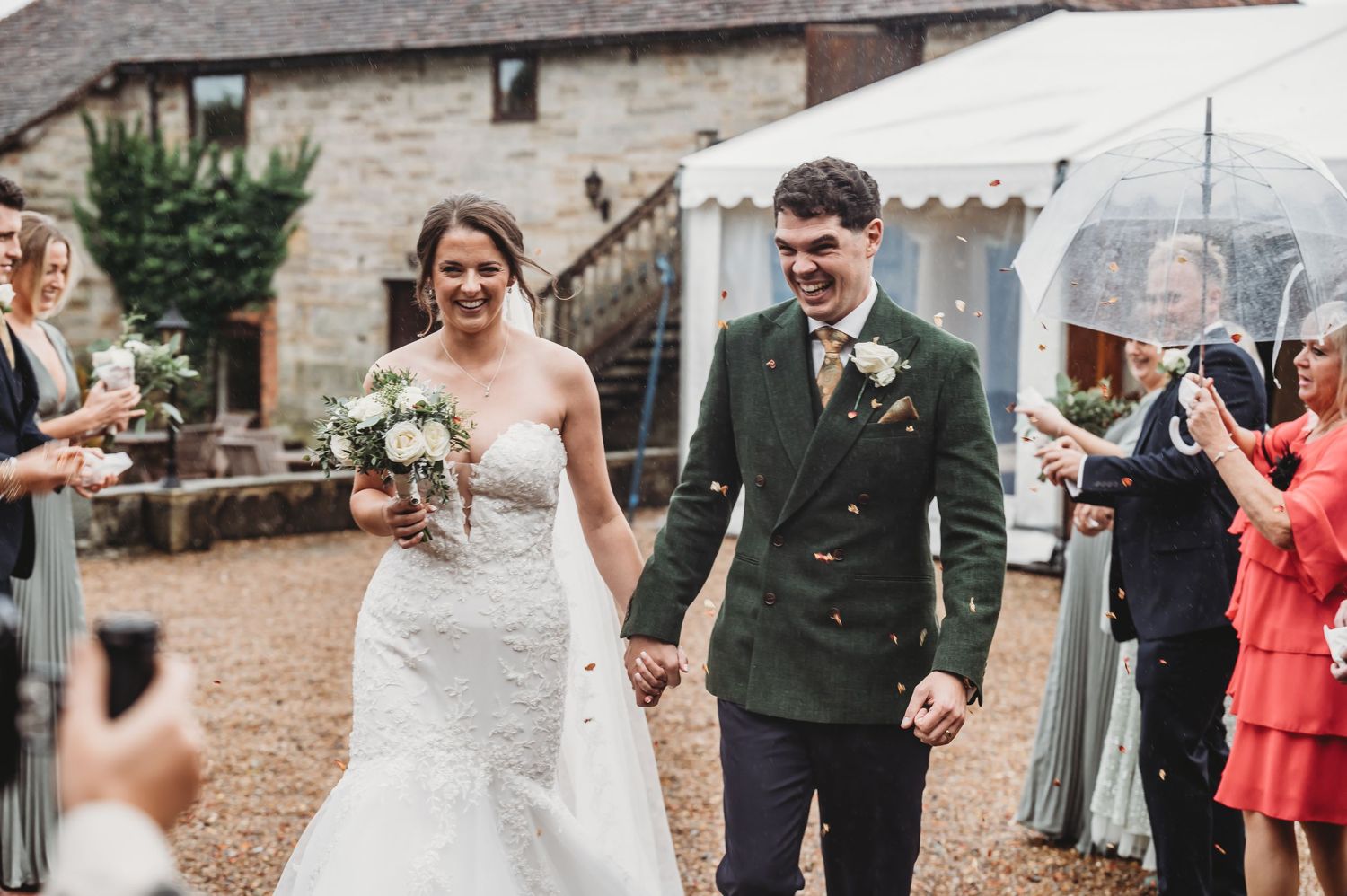 A newlywed couple walks down a gravel path while guests celebrate with umbrellas outside a rustic stone venue.