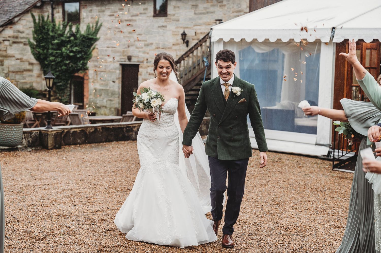Joyful couple walks down a gravel path while wedding guests celebrate their exit with confetti.