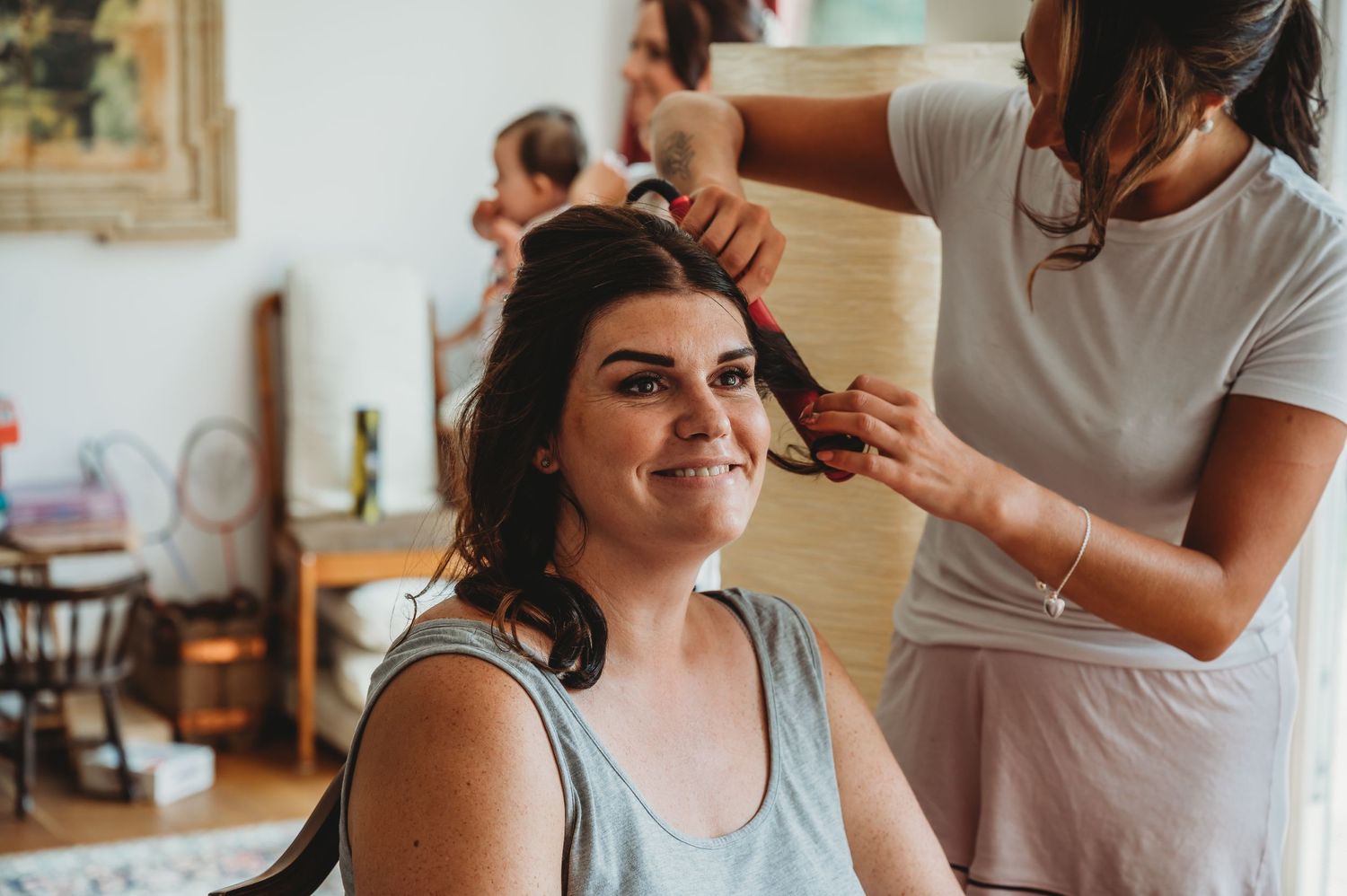 A hairstylist styles someone's dark curly hair in a salon interior with warm lighting and blurred background decor.