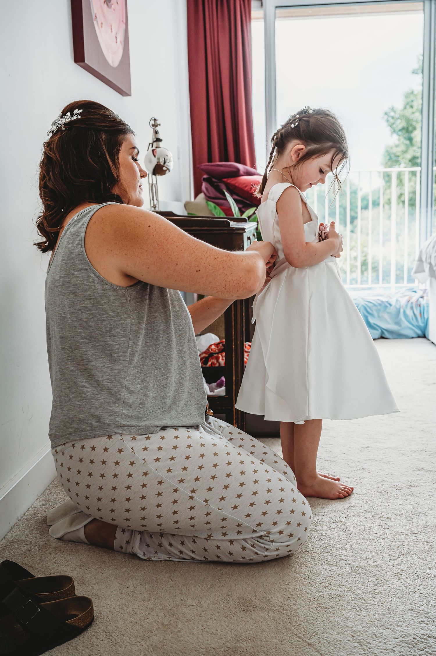 A tender moment shows someone helping adjust a child's white dress in a bedroom with red curtains and natural light.
