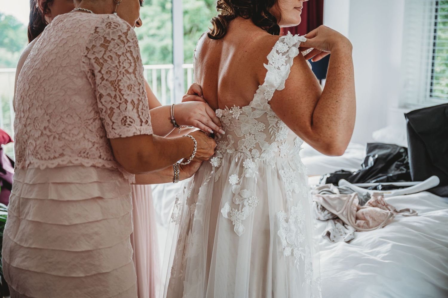 Detailed lace wedding dress being adjusted in bright dressing room with soft natural light.