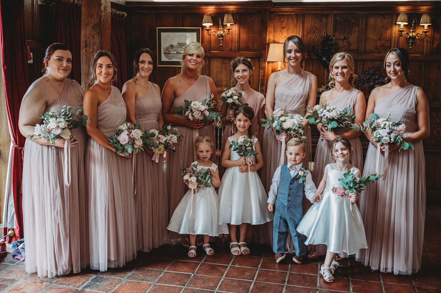 An elegant wedding party in blush pink bridesmaid dresses poses with flower girls in white and ring bearer in blue suit.