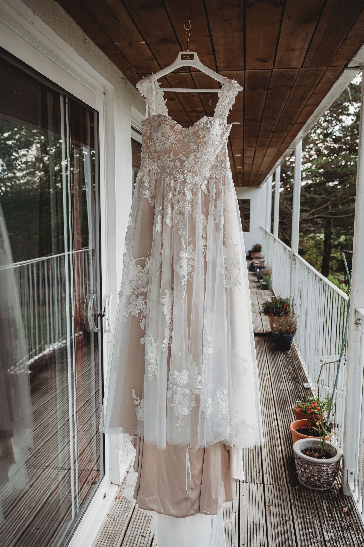 A delicate lace wedding dress hangs on a wooden porch with potted plants, featuring floral details and sheer overlay.