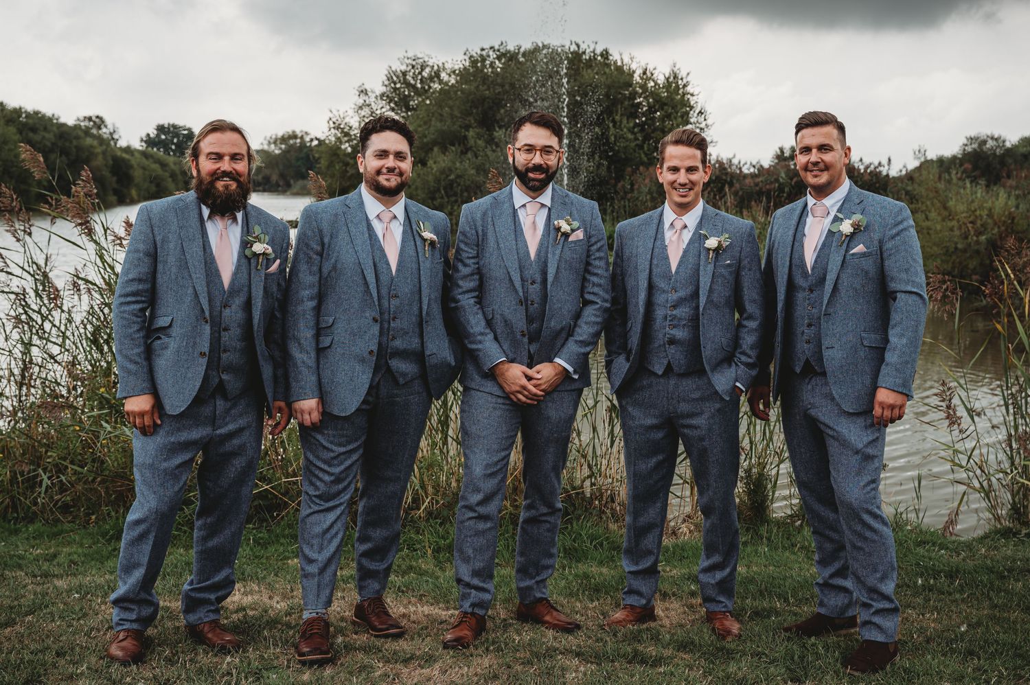 Five groomsmen wearing matching blue-gray suits pose together beside a pond on a cloudy day.