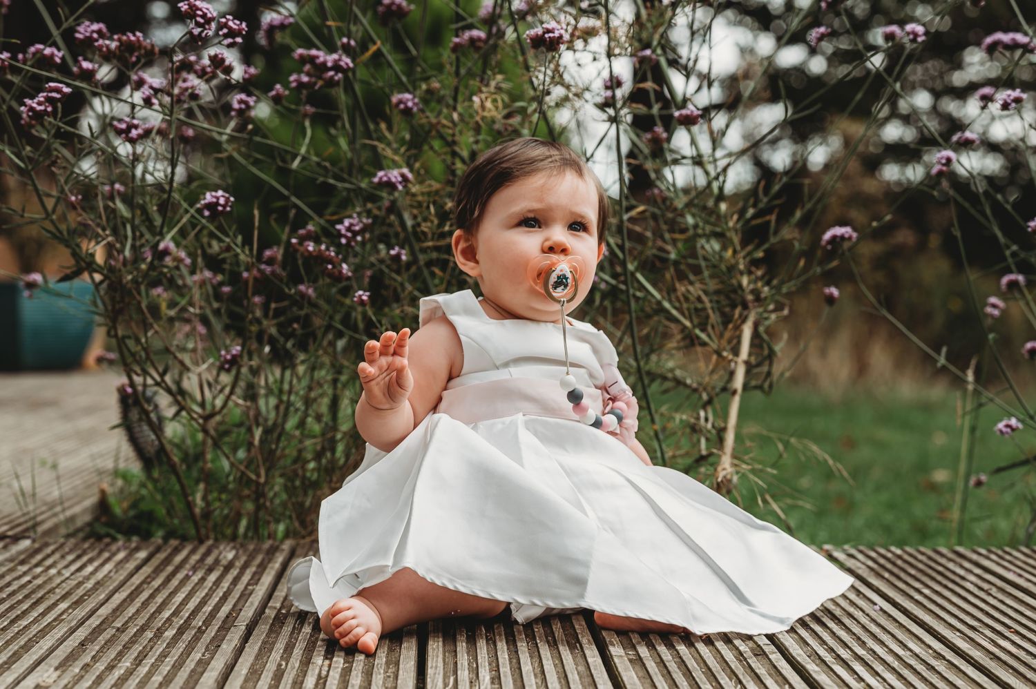 A small child wearing a white dress sits on a wooden deck surrounded by cherry blossoms in spring.