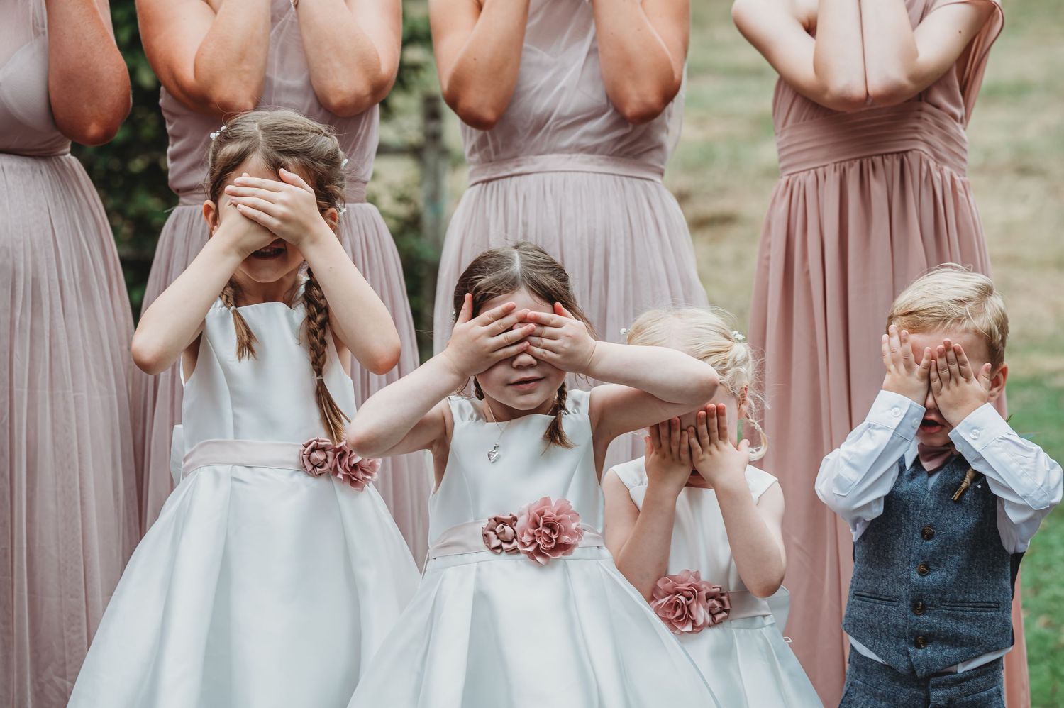 Children in formal wedding attire playfully cover their eyes while standing together at a celebration.