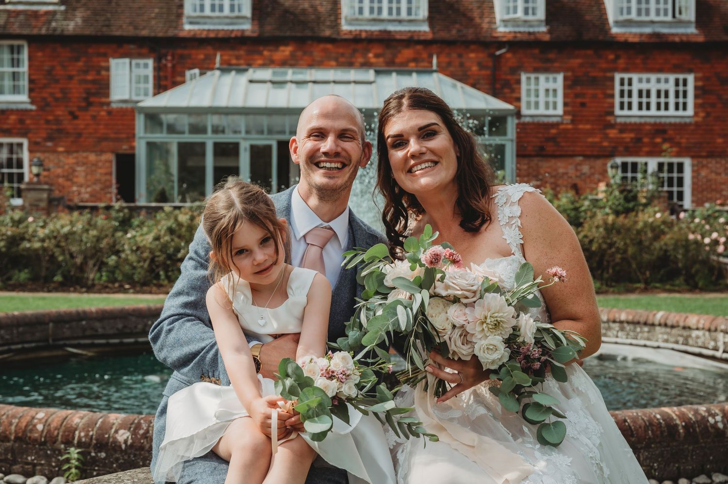 A wedding couple and child sit together by a brick mansion's fountain, surrounded by summer blooms and greenery.