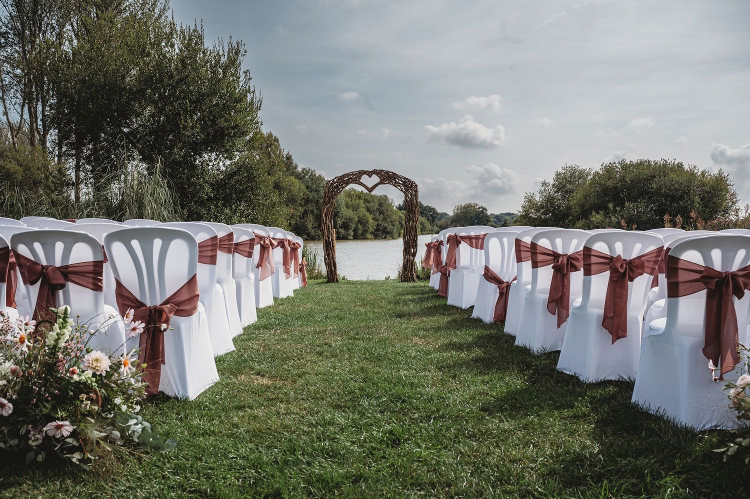 White wedding ceremony chairs with burgundy sashes line a grassy outdoor aisle leading to an arch.