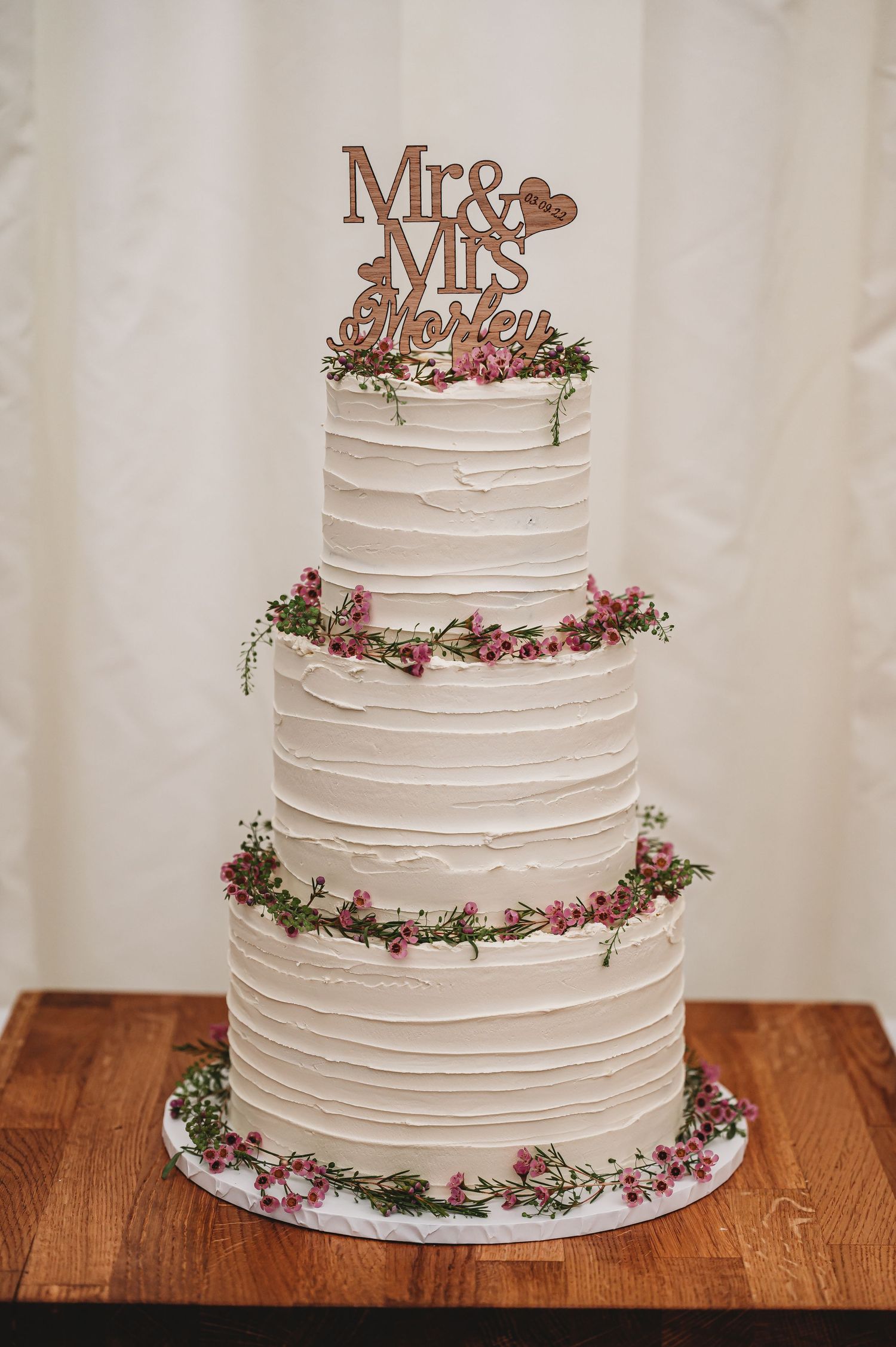Three-tier white wedding cake with textured frosting, Mr & Mrs topper, and delicate pink floral accents on wooden table.