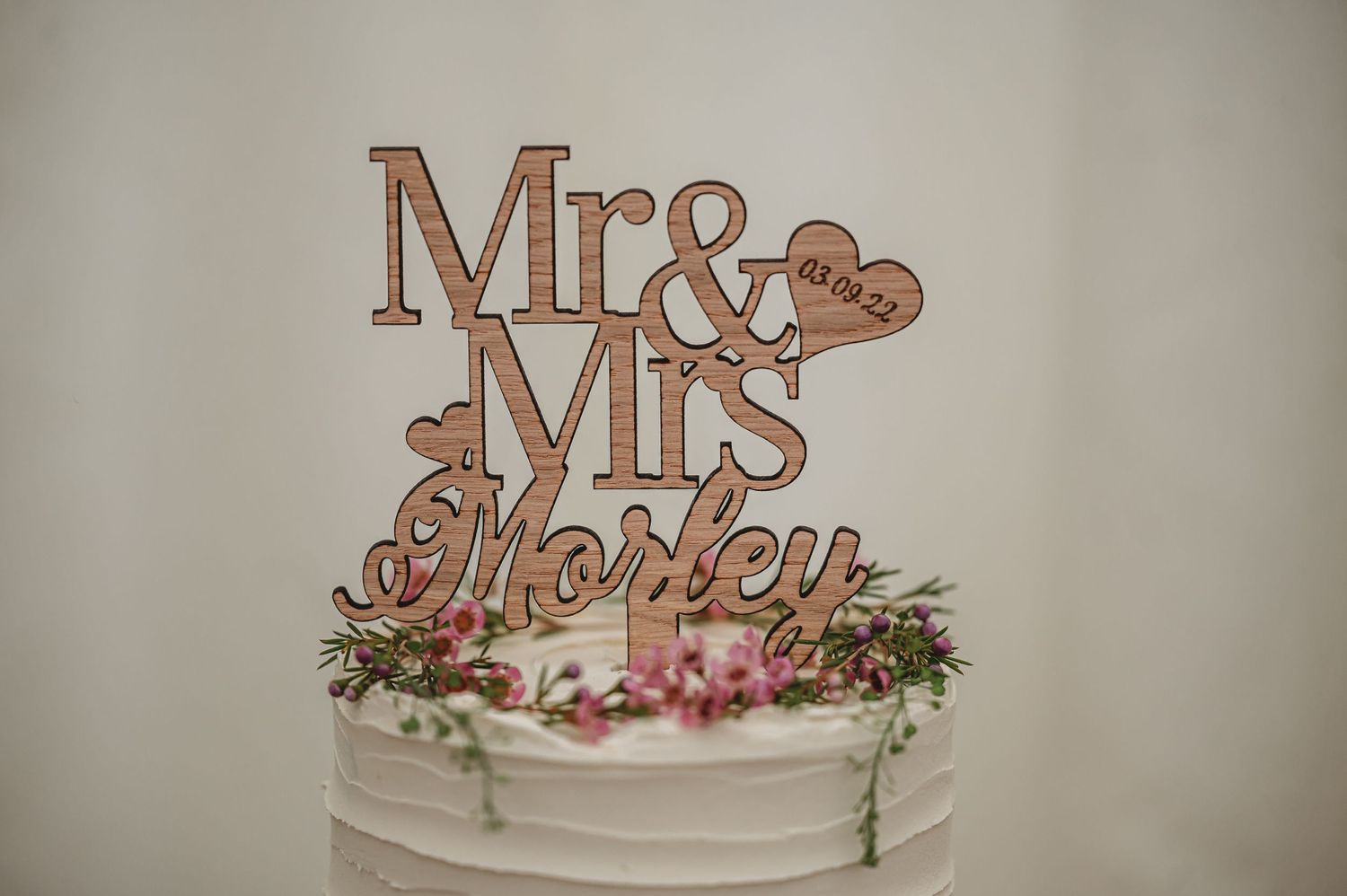 Wooden Mr & Mrs cake topper adorned with pink flowers and greenery on top of a white wedding cake.