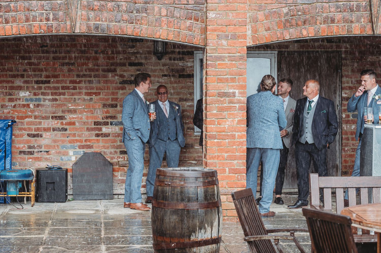 People gather and converse in small groups under brick archways near wooden barrels at a rustic venue.
