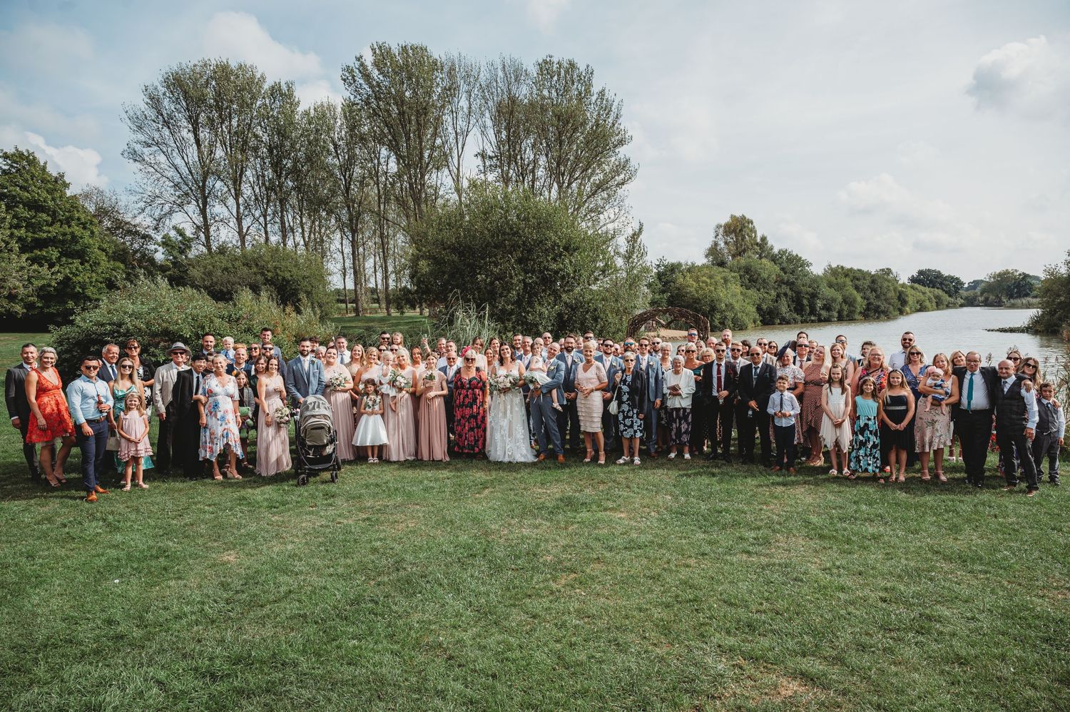 Large wedding party gathered on grass lawn near water and trees during outdoor celebration.