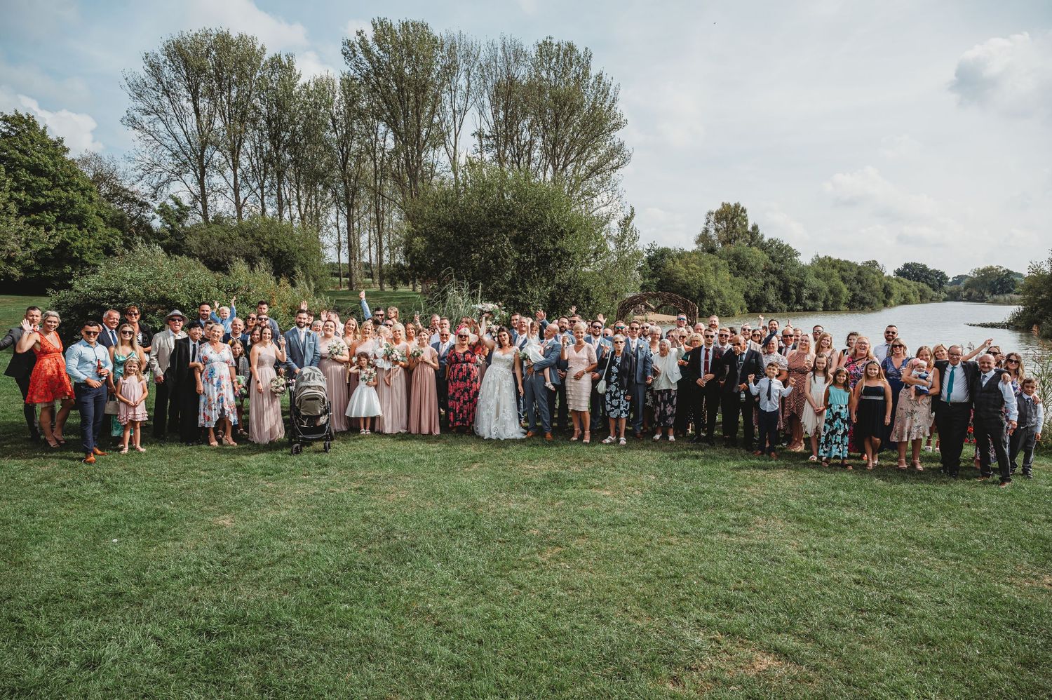 Large wedding party gathers for group photo on grassy lawn beside a lake with trees in background.