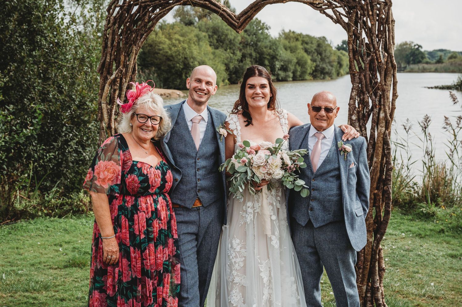 A group stands together under a rustic wooden arch by a serene lake during an outdoor celebration.