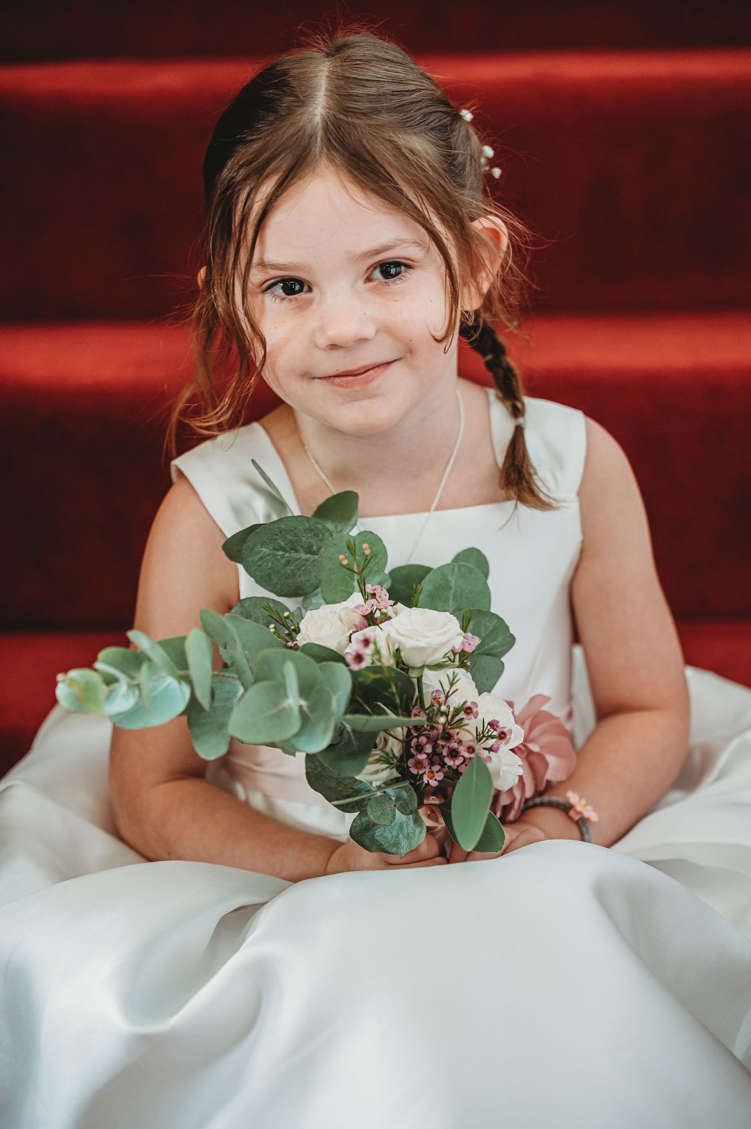 A small child in a white formal dress holds a delicate bouquet with eucalyptus leaves on red carpeted stairs.