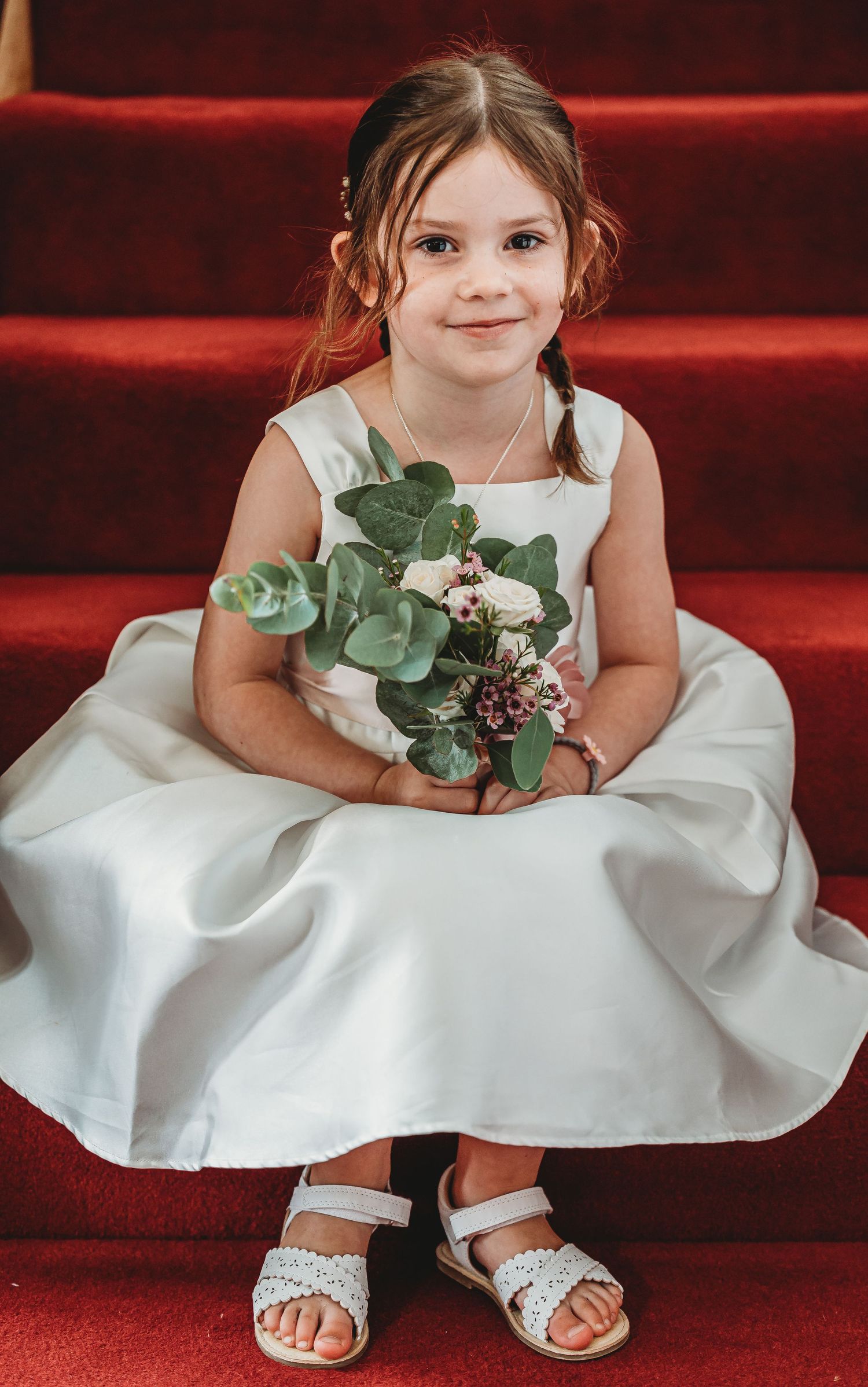 A young attendant in a white dress sits on red carpeted stairs holding a eucalyptus bouquet.