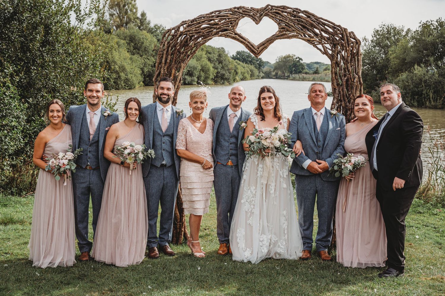 Wedding party poses together under rustic heart-shaped wooden arch in outdoor garden setting.