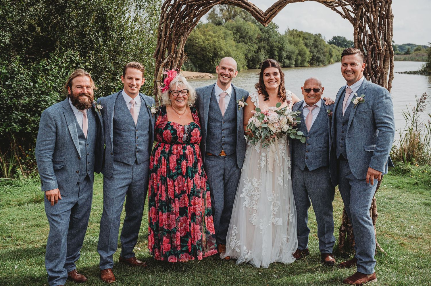 A group in formal attire poses outdoors by a lake with trees in the background during a wedding celebration.