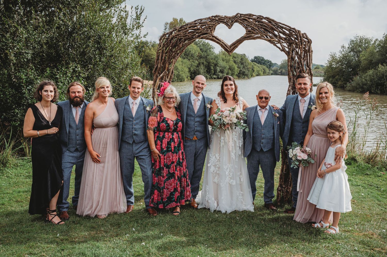 Wedding party poses under rustic heart-shaped arch by a lake during outdoor countryside celebration.
