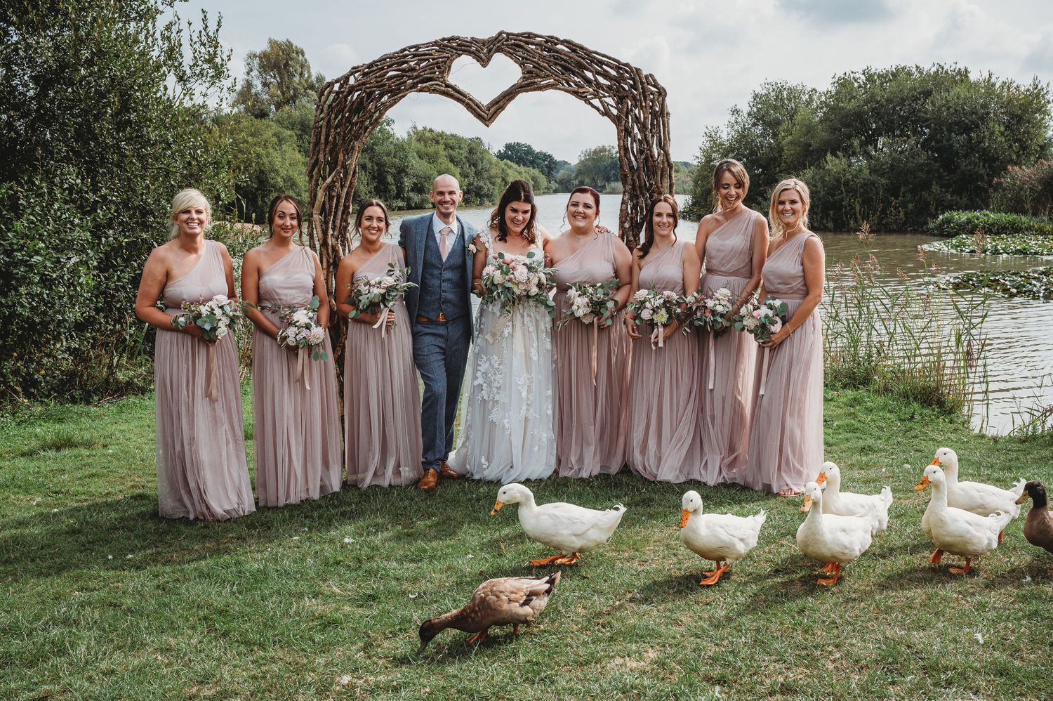 A wedding party poses outdoors under a heart-shaped arch with white geese wandering in the foreground by a pond.