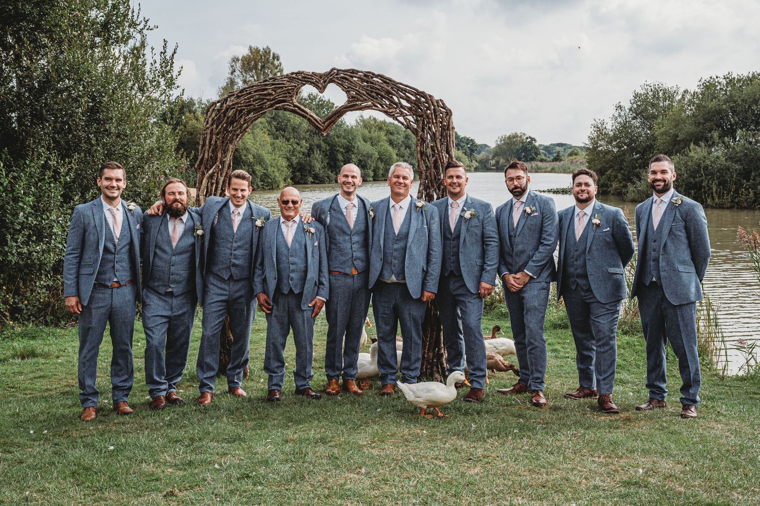 Wedding party in matching gray suits pose in front of rustic wooden arch at outdoor ceremony.