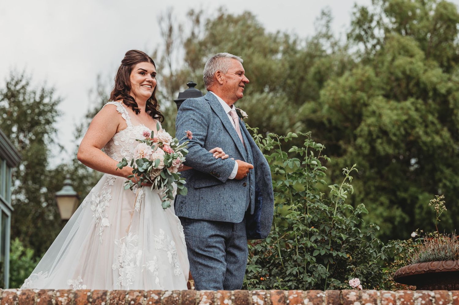A bride in a lace wedding dress walks arm in arm with someone in a blue suit on an outdoor path.