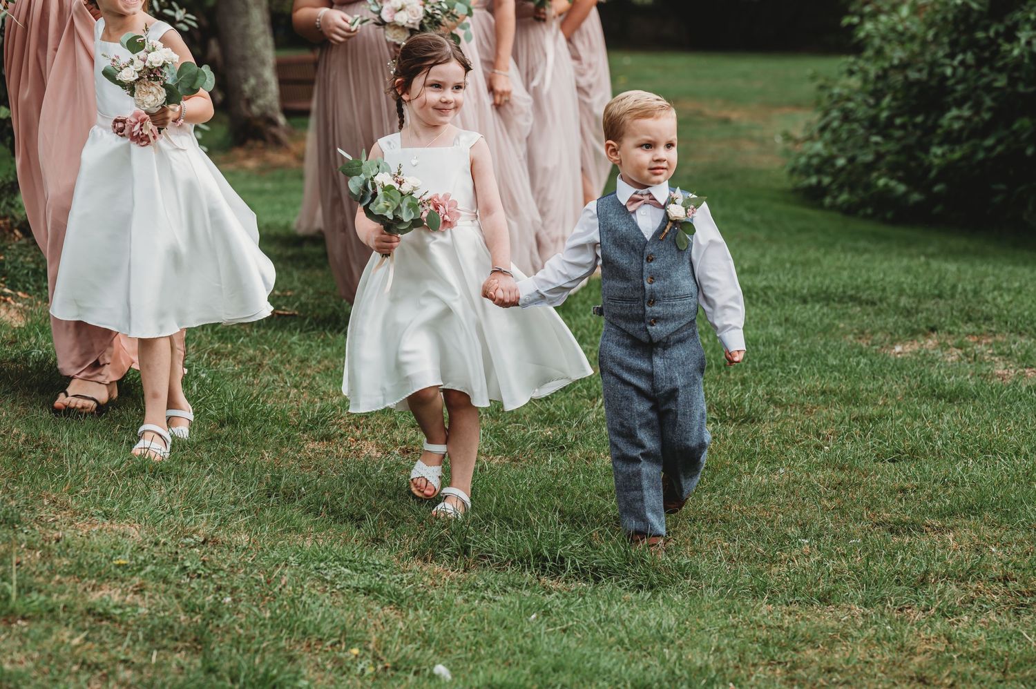Young participants in formal wedding attire walk together on a grassy lawn during an outdoor ceremony.