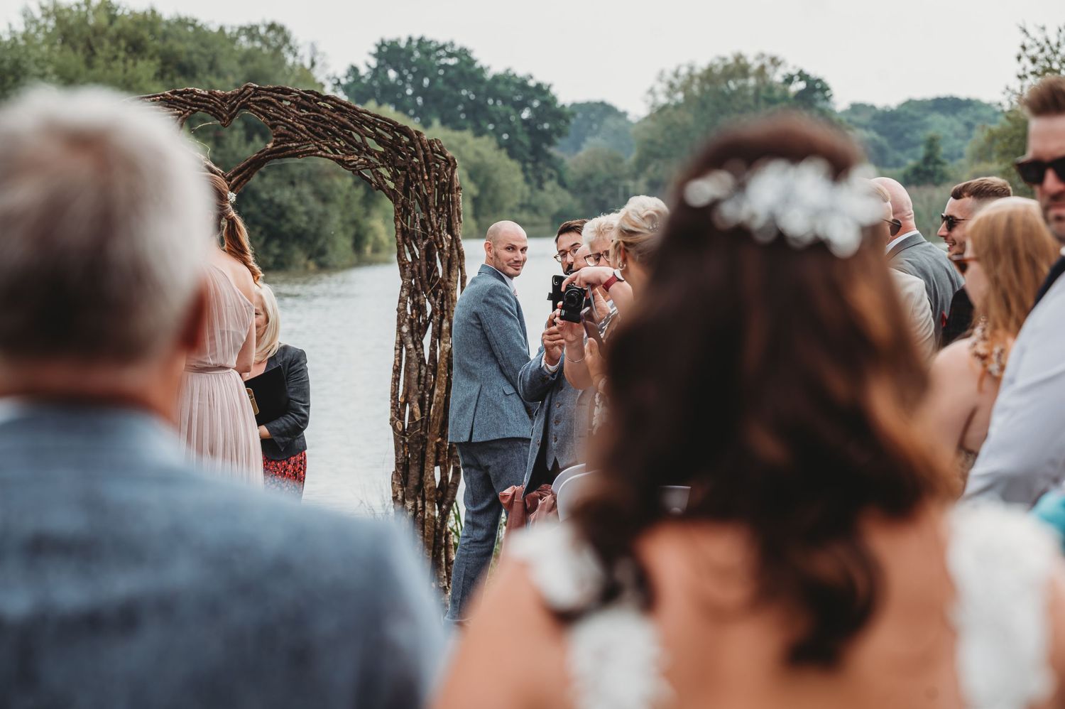 A rustic outdoor wedding ceremony takes place under a wooden arch by a lake on a late summer afternoon.