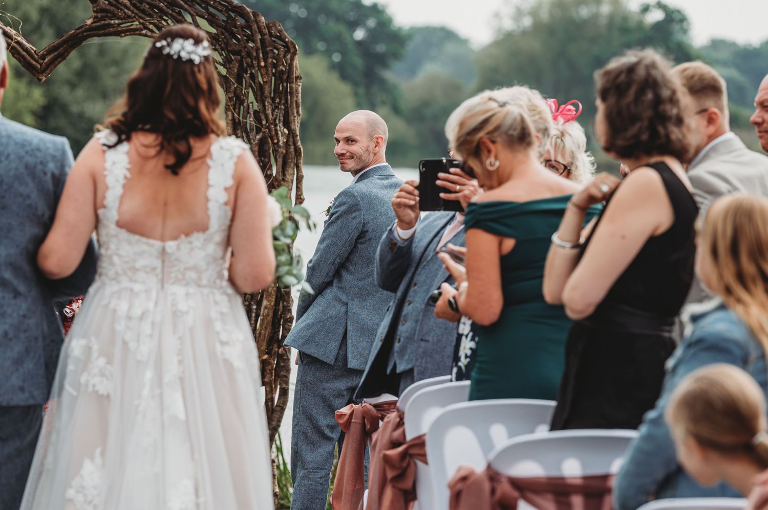 Wedding guests mingle at an outdoor reception under a tree while enjoying drinks and taking photos.