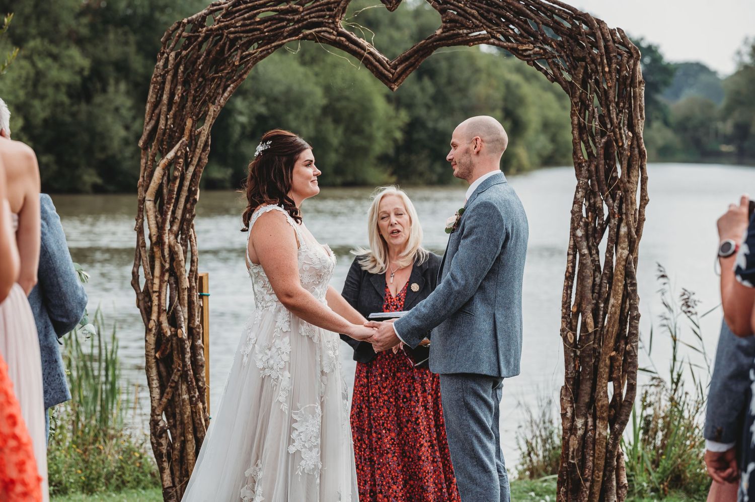 Outdoor wedding ceremony under rustic twig arch by serene lakeside with officiant and wedding party.