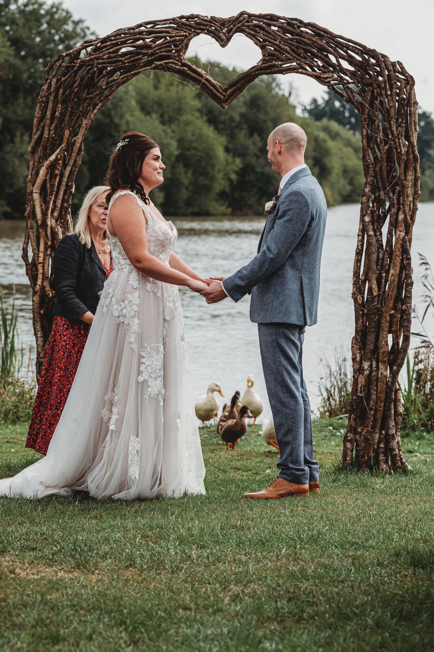 Rustic wedding ceremony under heart-shaped twig arch by lakeside with geese wandering in background.
