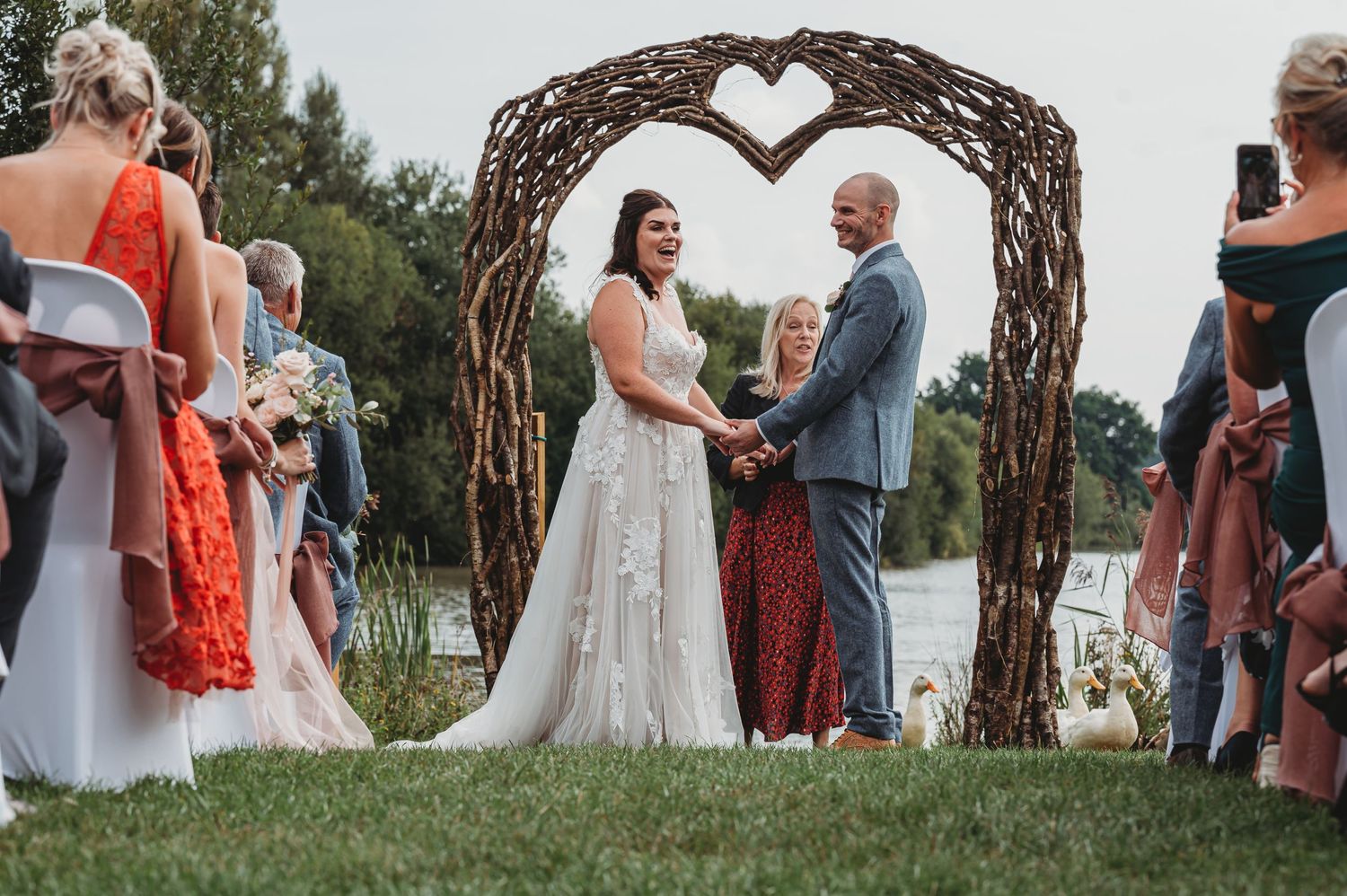 An outdoor garden wedding ceremony takes place under a rustic heart-shaped arch made of twigs on a grassy lawn.