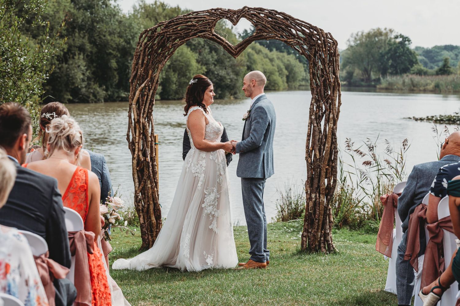 Outdoor wedding ceremony under rustic wooden arch with heart detail beside serene lake on sunny day.