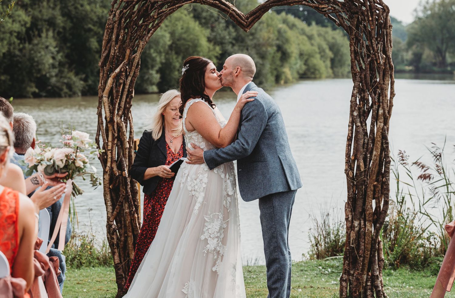 Wedding couple shares a kiss under rustic wooden arch by lakeside with guests watching in orange dresses.