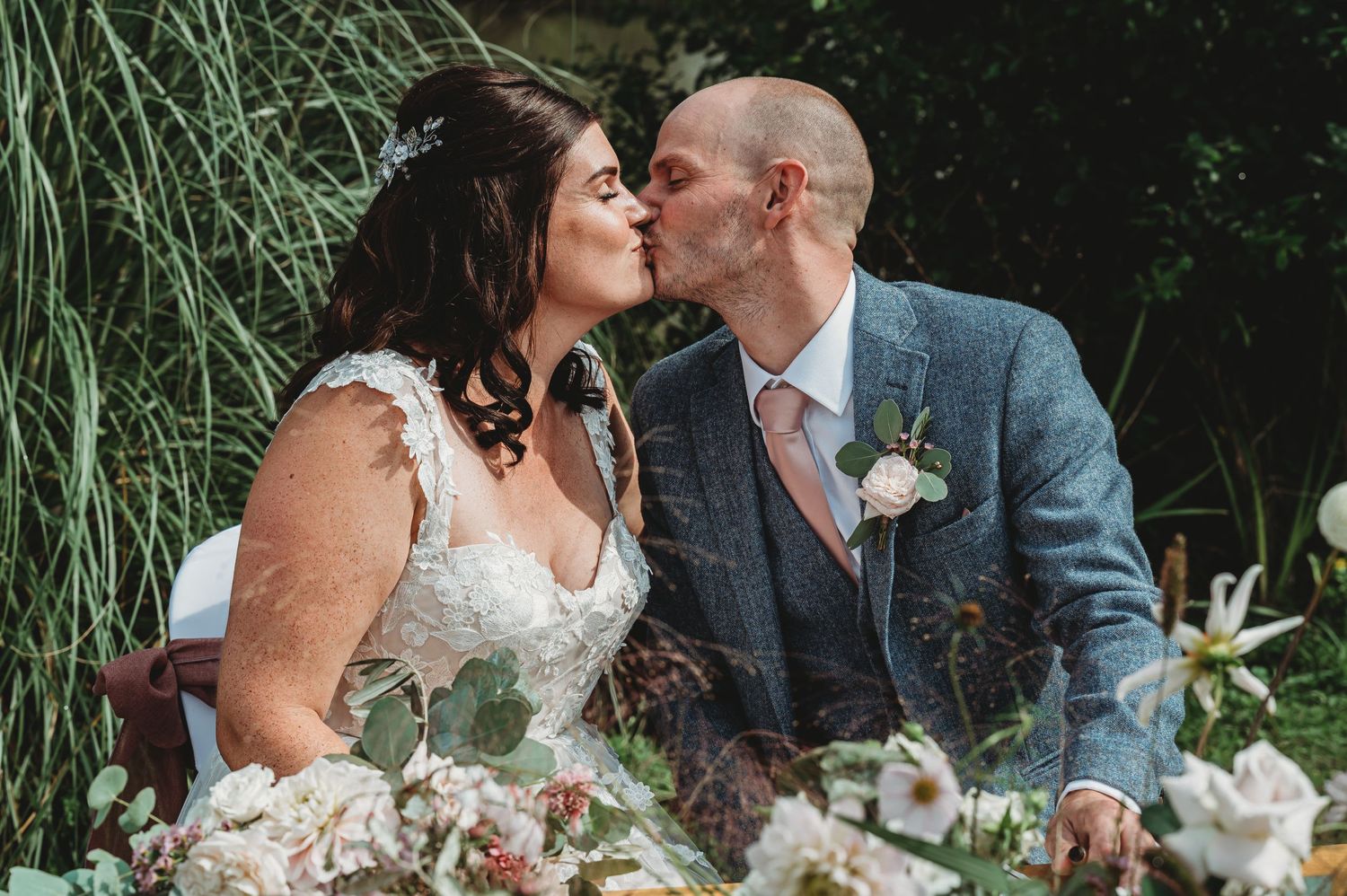 A romantic kiss under greenery at an outdoor wedding celebration with soft pink and white floral decorations.