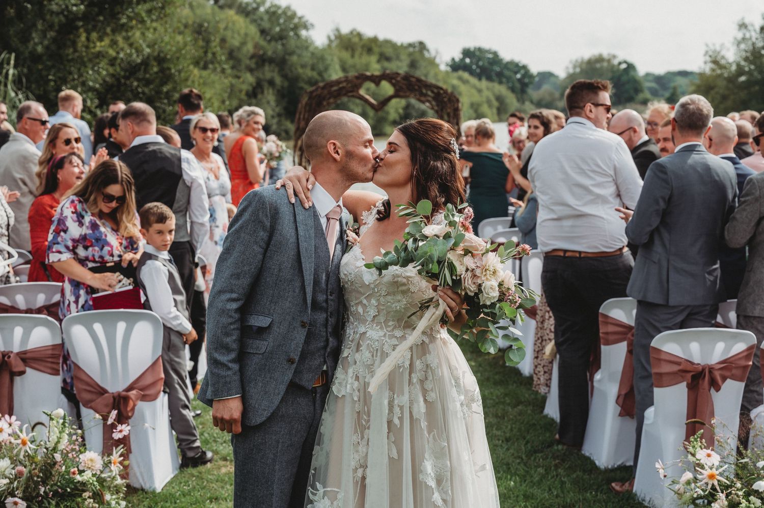 Outdoor wedding celebration with white chairs set up on grass as a couple shares a moment amid gathered guests.
