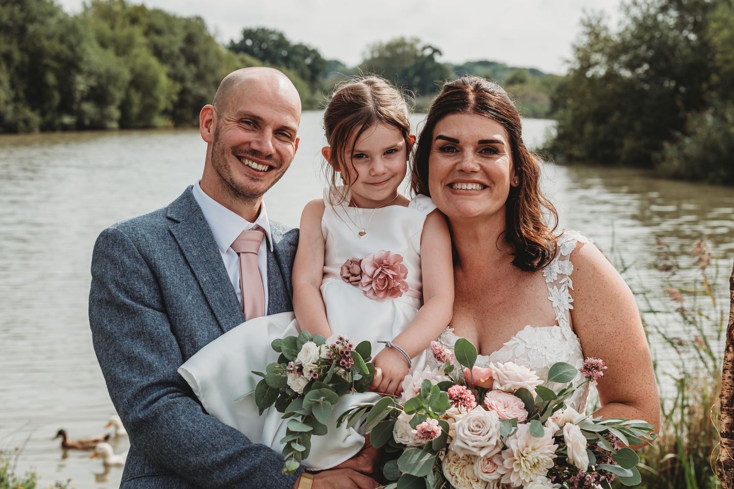 Outdoor wedding portrait by a lake with a beautiful floral bouquet and ducks swimming in the background.