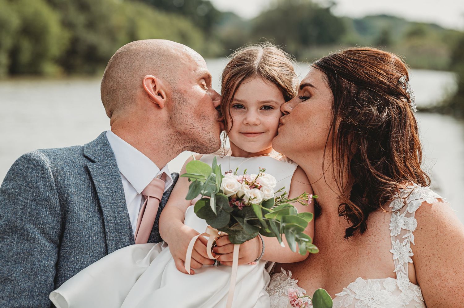 A romantic wedding moment by a lake with the couple embracing while holding a delicate white floral bouquet.