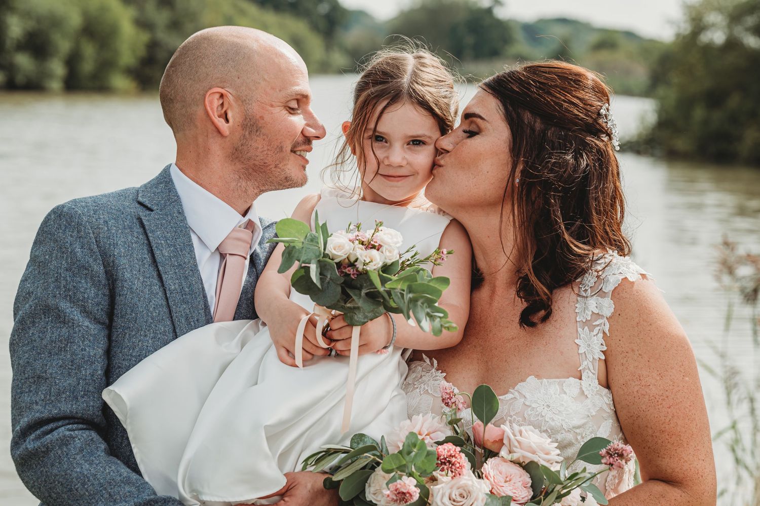 A wedding couple and small child hold pink and white floral bouquets while posing near a waterfront on their special day.