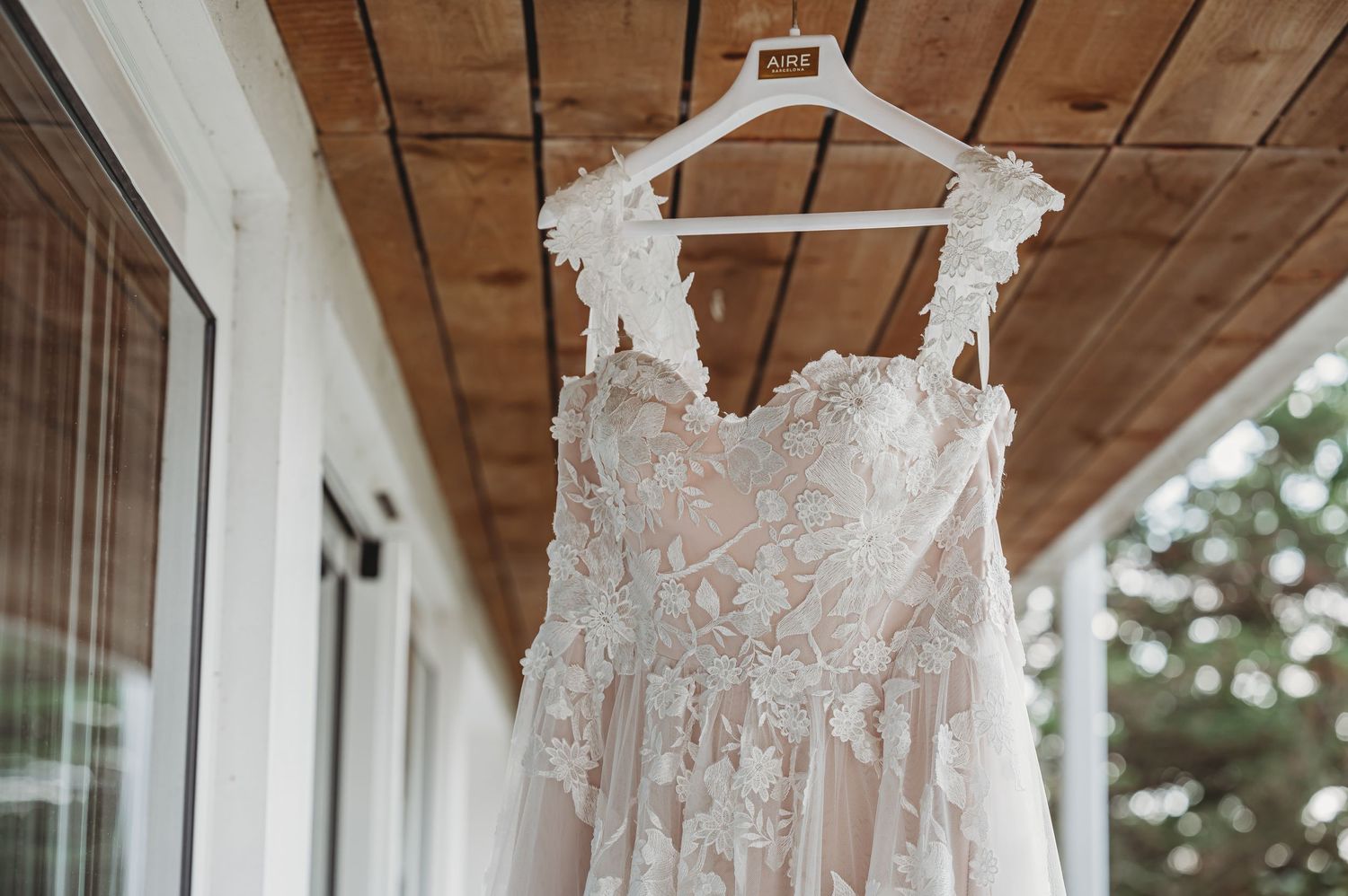 A delicate lace wedding dress with floral appliques hangs on a white hanger against a wooden porch ceiling.