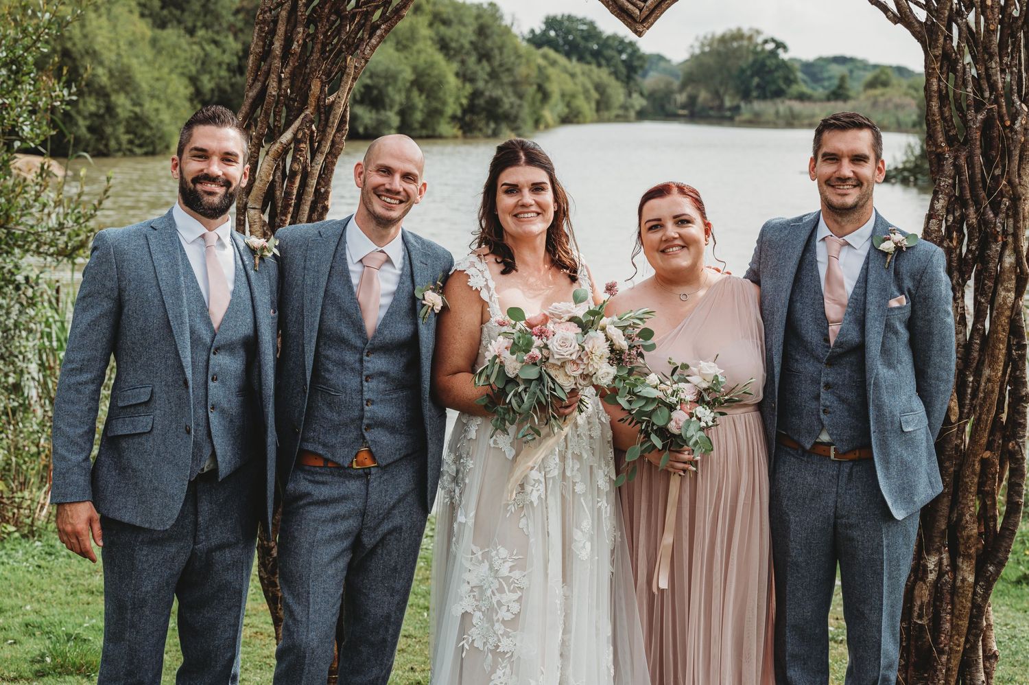 A wedding party in blue-gray suits and soft blush gowns poses together outdoors by a lake under trees.